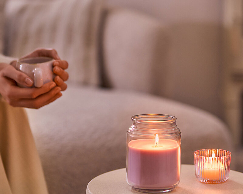Person hält eine Tasse Kaffee auf einem gemütlichen Sofa, im Vordergrund brennt das True Scents Duftglas in Lavendel neben einer kleinen Teelichthalter-Kerze auf einem runden Beistelltisch