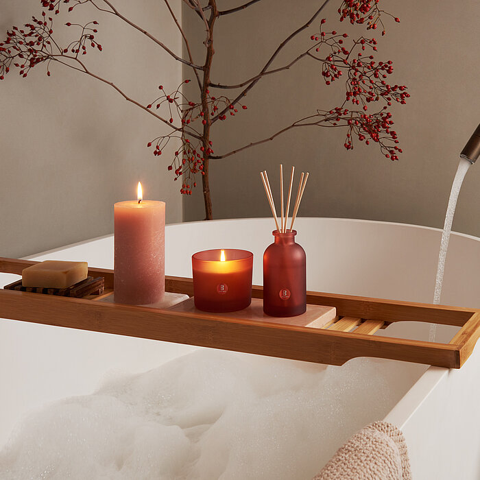 Eine Badewanne mit Wellness-Stimmung. Über der Wanne steht ein Holzbrett mit True Joy Duftglas und Raumduft in Rosa, sowie eine rosa Rustik Stumpenkerze von Bolsius.