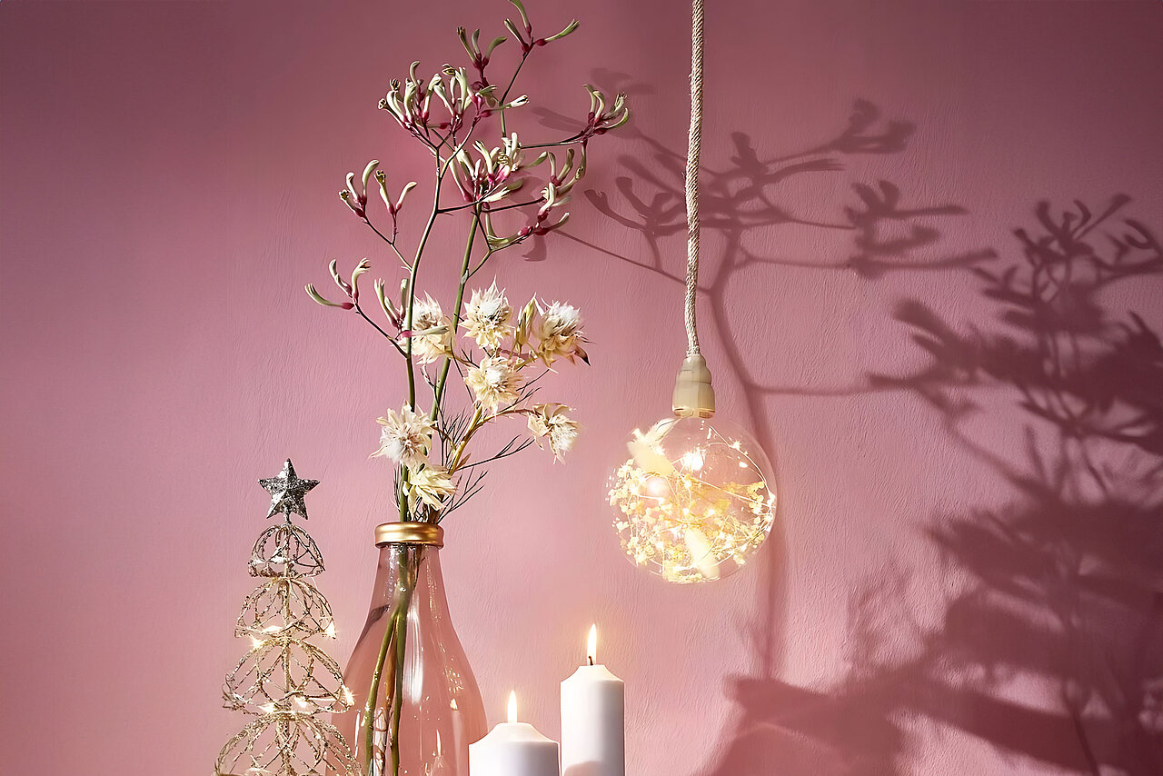 Vor einer altrosa Wand stehen verschiedene Dekorationsgegenstände, die einladend wirken: Ein filigraner Tannenbaum aus goldenem Draht, eine transparente Vase mit blühenden Zweigen, eine dekorative Glühbirne mit LED-Kette und getrockneten Blüten sowie zwei weiße Stumpenkerzen, die eine gemütliche Atmosphäre schaffen. Die Dekoobjekte werfen sanfte Schaffen auf die Wand.