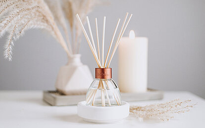 Ein freundlicher, heller Raum mit einer hellgrauen Wand. Auf einem weißen Tisch stehen Schilf in einer weißen Vase und eine cremefarbene Stumpenkerze im Hintergrund. Im Vordergrund ist ein eleganter Raumduft Diffuser mit Rattanstäbchen zu sehen. Dieser läßt sich als Alternative zu Duftkerzen einsetzen.