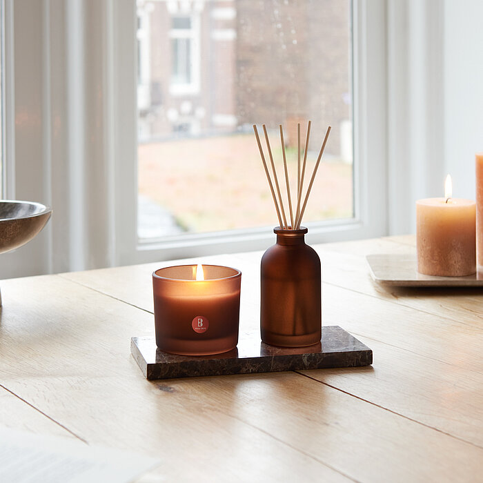 Ein heller, freundlicher Raum mit Duftglas und Raumduft-Diffuser auf dem Tisch. Im Hintergrund brennen Bolsius Rustik-Kerzen.