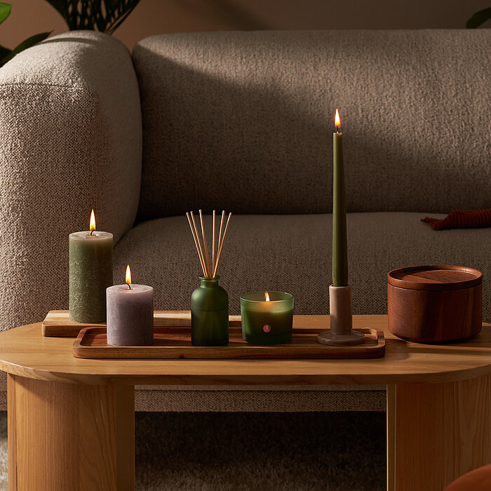 Ein gemütliches Wohnzimmer mit brennden Kerzen auf einem Couchtisch. Zwischen den Rustik- und Tafelkerzen stehen Raumduft Diffuser und Duftglas in Grün aus der True Joy Kollektion von Bolsius.