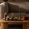 Ein gemütliches Wohnzimmer mit brennden Kerzen auf einem Couchtisch. Zwischen den Rustik- und Tafelkerzen stehen Raumduft Diffuser und Duftglas in Grün aus der True Joy Kollektion von Bolsius.