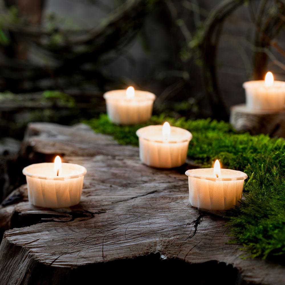 Nahaufnahme einer brennenden Stearin Kerze in einem orangefarbenen Glas. Im Hintergrund erkennt man noch zwei weitere Kerzen, eine mit grünem Glas und eine mit klarem. Die Gläser stehen auf dichtem, grünem Moos.
