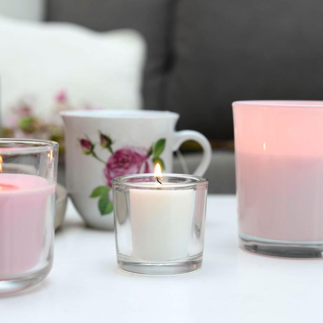 Romantisch gedeckter Tisch mit einer Porzellantasse mit Rosenmotiv. Im Mittelpunkt steht eine weiße, brennende Votivkerze in einem durchsichtigen, dicken Glashalter. Daneben stehen zwei weitere brennende rosa Kerzen.