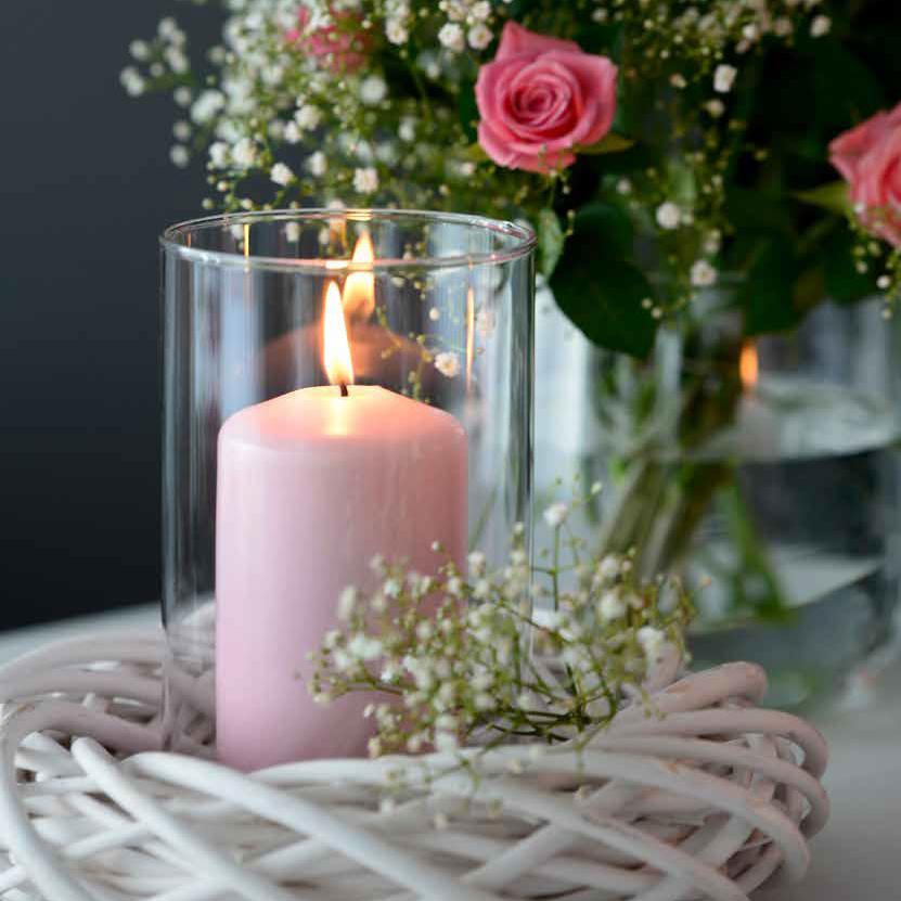 Ein geflochtener, weißer Weidekranz mit einer hohen Glas Vase und einer brennenden, rosa Stumpenkerze in der Mitte. Im Hintergrund erkennt man einen Strauß rosa Rosen mit Schleierkraut dazwischen. Neben dem Kerzenglas liegt ein weiterer Zweig Schleierkraut.