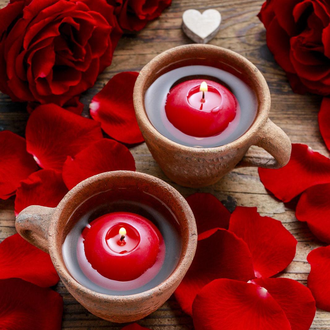 Zwei romantische Kerzen in Form von roten Schwimmkerzen, die jeweils in einer rustikalen Tasse aus Ton schwimmen. Die Tassen stehen auf einem groben Holztisch zwischen roten Rosen, Blüten und kleinen, weißen Holzherzen.