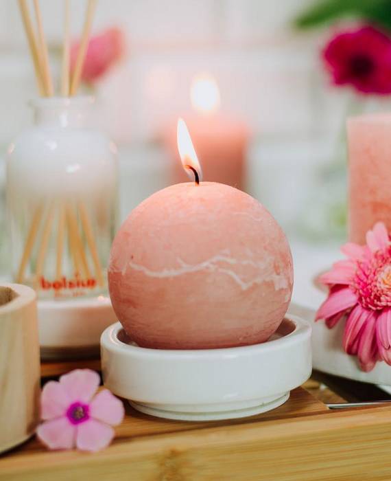 Eine brennende Rustik Kerze und eine Stumpenkerze in zartem Rosa auf weißen Kerzenhaltern. Dazwischen liegen rosa Blüten, auf einem Tablett aus Holz, das über einer Badewanne steht.