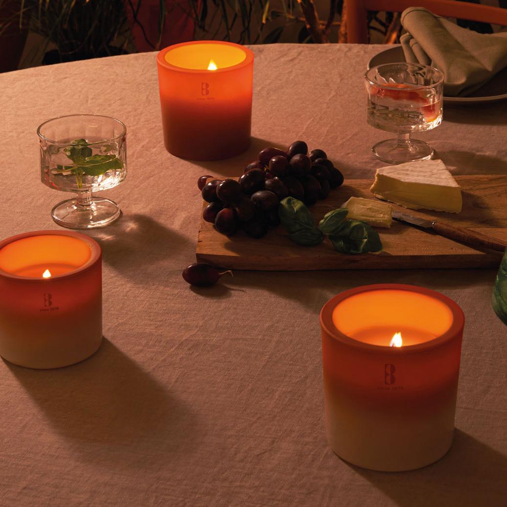 Drei brennende Outdoor Kerzen in orange auf einem gemütlich beleuchteten Tisch, auf dem außerdem ein Brett mit Käse und Trauben auf einer rustikalen Tischdecke stehen.