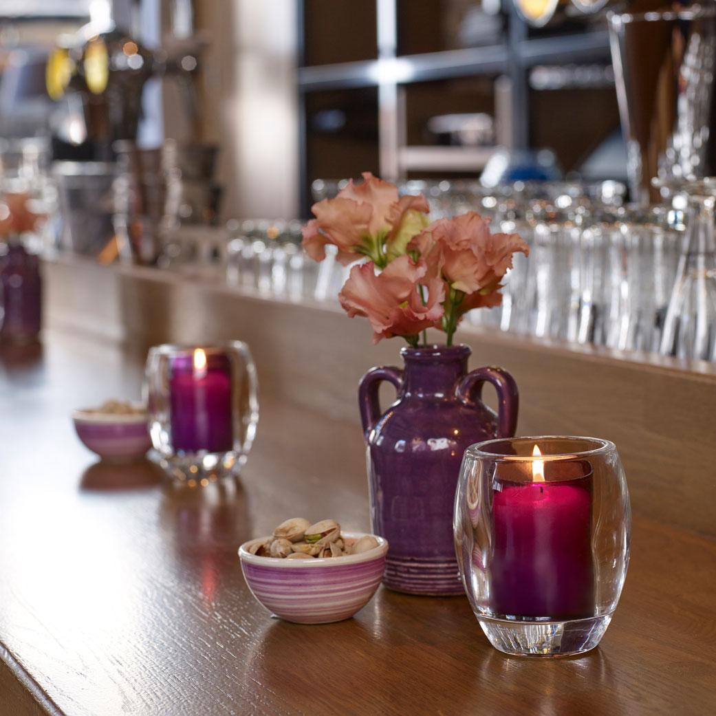 Ein Bartresen in einem Lokal, dekoriert mit violetten Keramikvasen und passenden Keramikschalen mit Pistazien. Daneben steht jeweils eine violette Relight Kerze in einem eleganten, ovalen Glashalter.