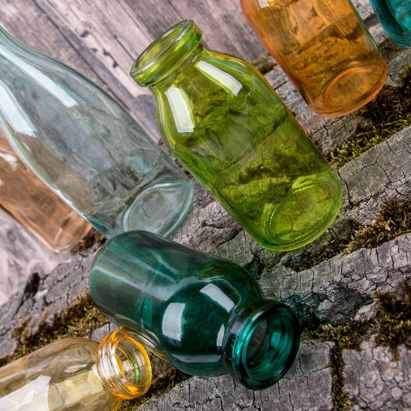 Eine Nahaufnahme verschiedener Deko-Flaschen in bunten Farben wie Grün, Gelb, Orange oder Hellblau. Die Flaschen stehen und liegen wild verteilt auf einem mit Moos bewachsenen Baumstamm.
