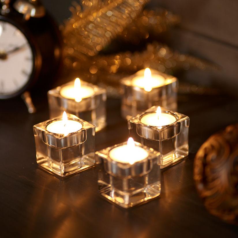 Fünf quadratische Glas-Teelichthalter mit brennenden Teelichtern darin, die für ein warmes, goldenes Licht sorgen. Mit ihnen auf dem Tisch steht ein alter Blechwecker und eine golden glitzernde Feder Dekoration.