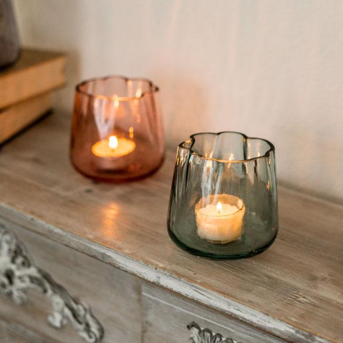 Drei bunte Teelichtgläser mit gewelltem Rand stehen auf einem antiken Schränkchen neben einer brennenden, cremefarbenen Stumpenkerze. In den Glashaltern brennen verschiedene Frühlingsdeko Duftteelichter.