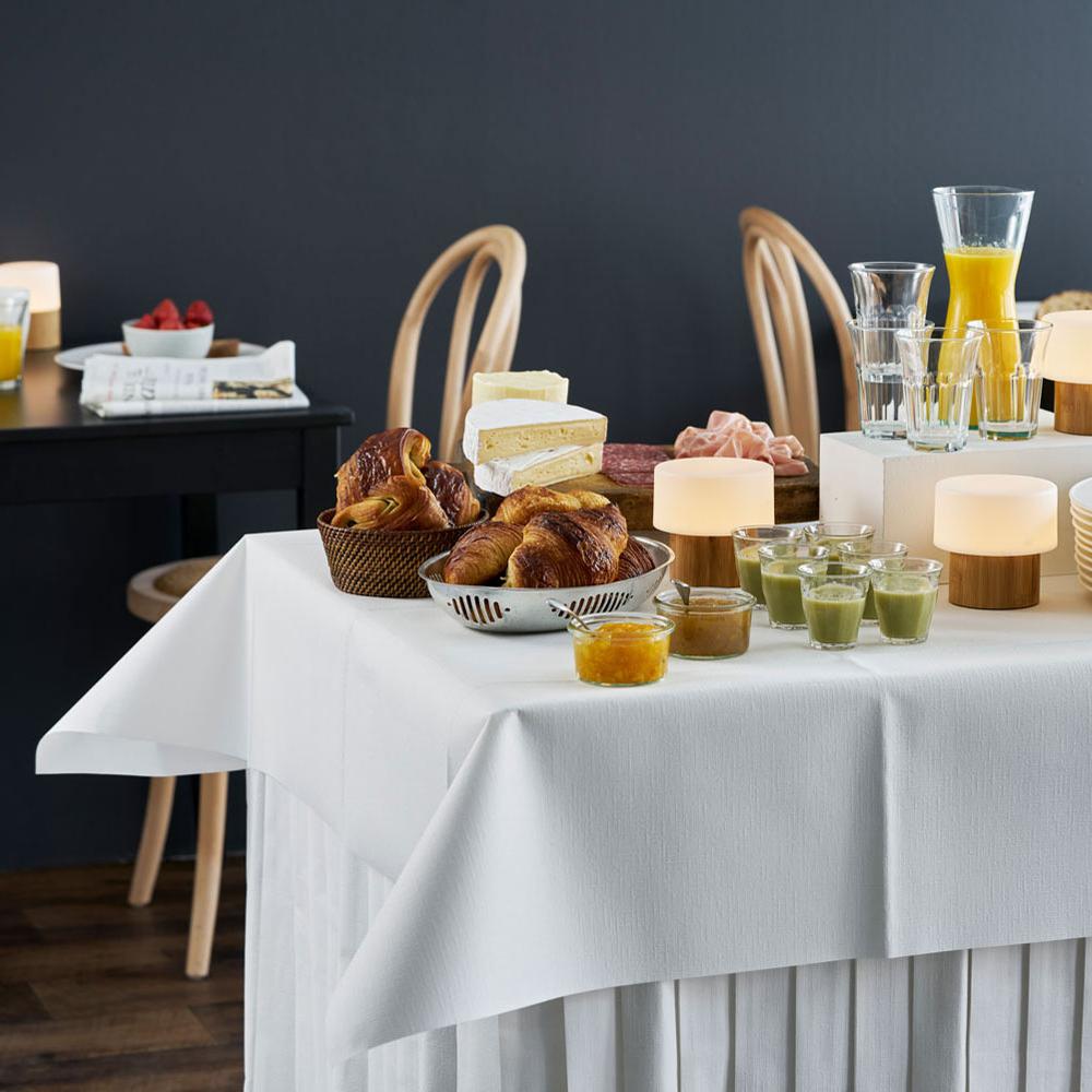 Ein großzügig bestücktes Frühstücksbuffet mit Croissants, Orangensaft, Müsli und Geschirr steht in einem Gastraum. Im Hintergrund sind gedeckte Frühstückstische zu sehen. Auf dem Tisch im Vordergrund liegt eine weiße Tischdecke.