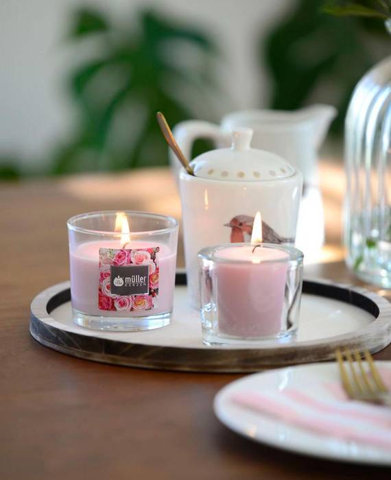 Romantisch gedeckter Tisch mit Porzellangeschirr mit verspieltem Rosen- und Amselmotiv und einer großen Glasvase mit Rosen im Hintergrund. Auf dem Tisch steht auch ein Tablett mit zwei brennenden Kerzen: eine Rosen-Duftkerze im Glas und eine rosa Votiv-Kerze in einem durchsichtigen Glashalter.