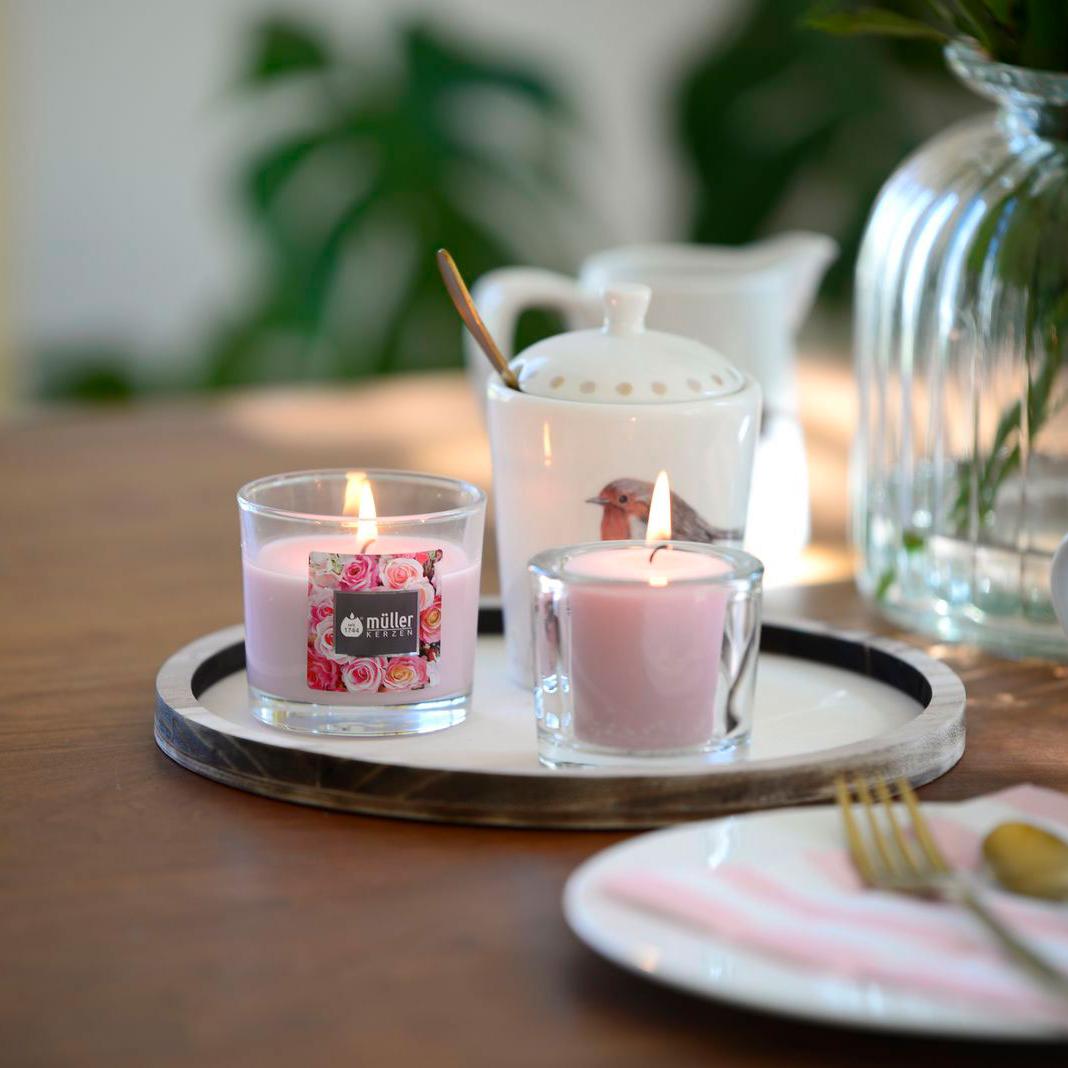 Romantisch gedeckter Tisch mit Porzellangeschirr mit verspieltem Rosen- und Amselmotiv und einer großen Glasvase mit Rosen im Hintergrund. Auf dem Tisch steht auch ein Tablett mit zwei brennenden Kerzen: eine Rosen-Duftkerze im Glas und eine rosa Votiv-Kerze in einem durchsichtigen Glashalter.