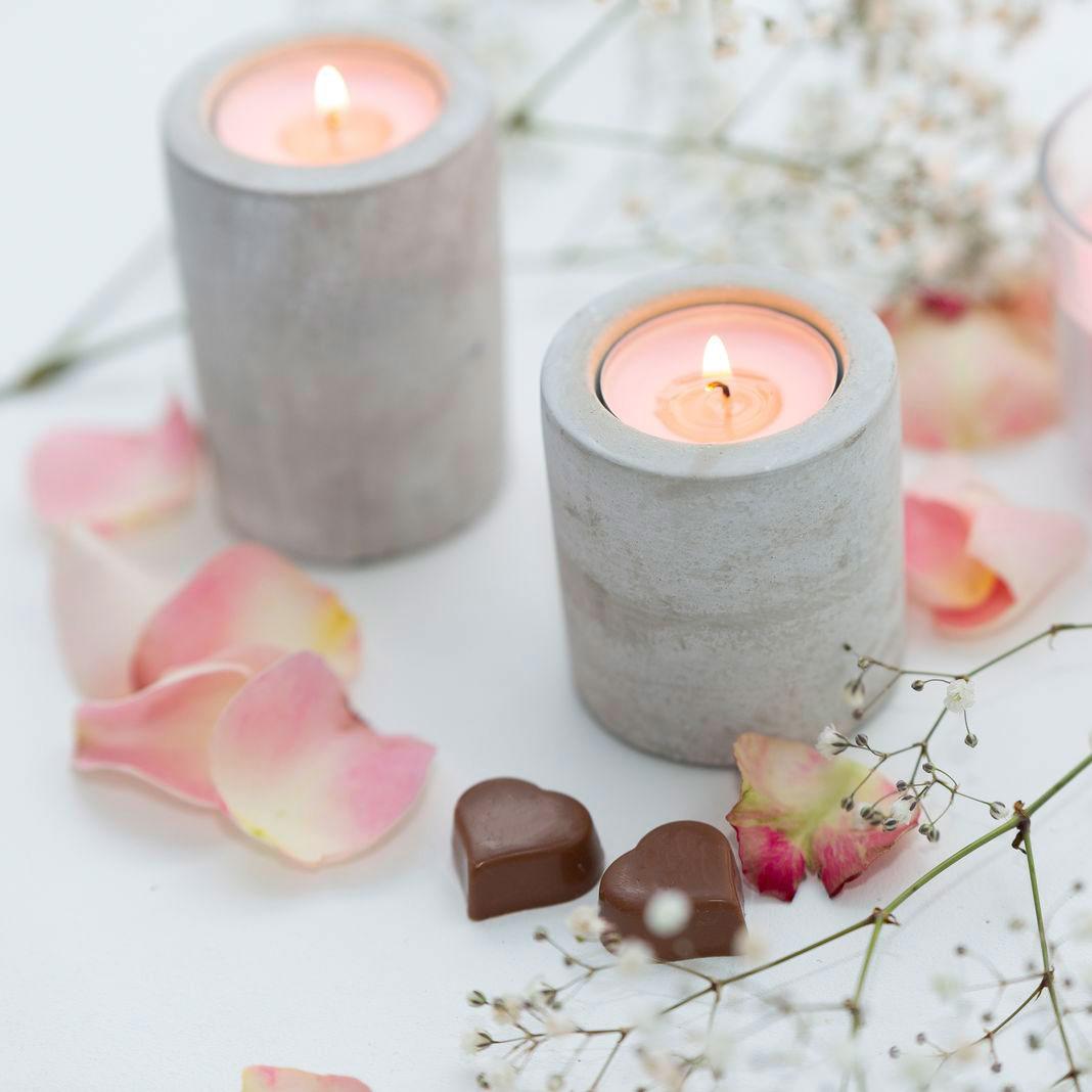 Ein weißer Untergrund, romantisch mit rosa und weißen Blüten und zwei Schoko-Herzen dekoriert. Dazwischen stehen eine brennende rosa Kerze im Glas, sowie zwei rosa Teelichter in hohen Beton-Kerzenhaltern.