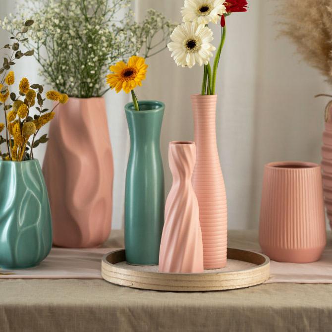 Ein Tisch, bedeckt mit braunen und rosafarbenen Leinentischdecken, auf dem viele verschiedene Deko Vasen aus Keramik stehen. Die Vasen in rosa und mintgrün haben verschiedene Formen, gewellte oder glatte Oberflächen und sind befüllt mit verschiedenen Blumen wie Gerbera, Schilf oder Schleierkraut.