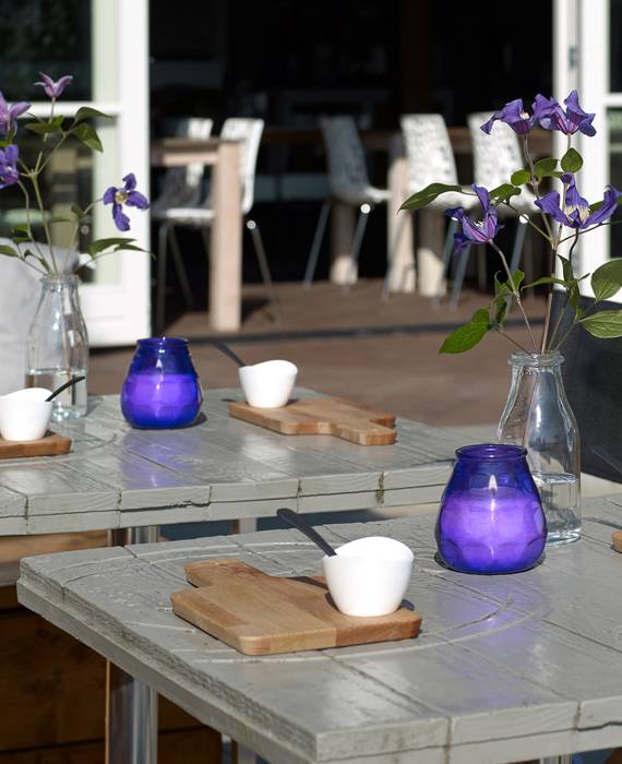 Zwei graue Terrassentische mit Frühstücksbrettchen und violetten Blumen in einer Glasvase. Auf jedem Tisch steht ein violettes, brennendes Windlicht.