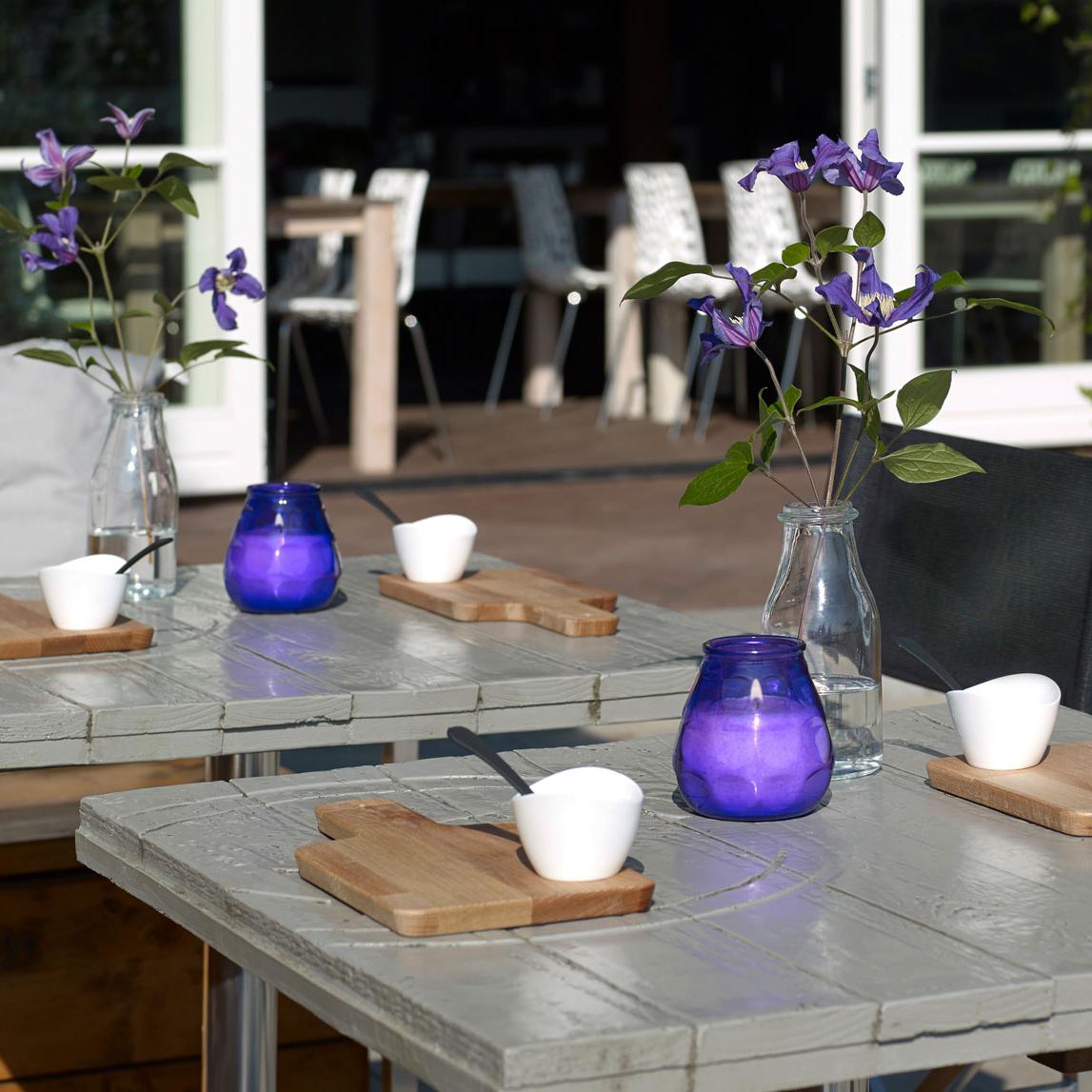 Zwei graue Terrassentische mit Frühstücksbrettchen und violetten Blumen in einer Glasvase. Auf jedem Tisch steht ein violettes, brennendes Windlicht.