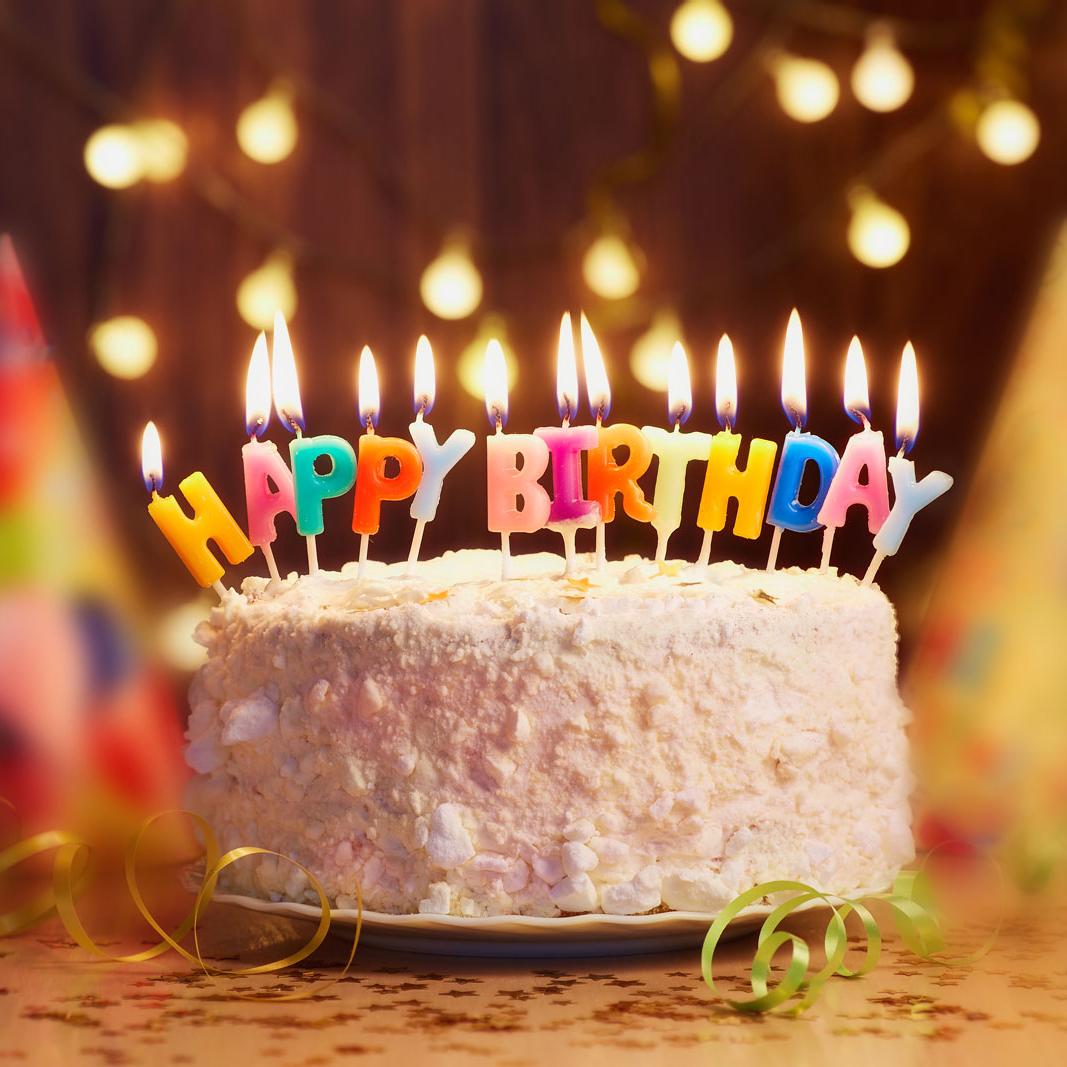 Nahaufnahme einer Geburtstagstorte mit weißem Überzug. Im Hintergrund sind bunte Partyhüte und Luftschlangen zu erkennen. Auf der Torte brennen in bunten Buchstaben kleine Tortenkerzen, die den Schriftzug “Happy Birthday” bilden.