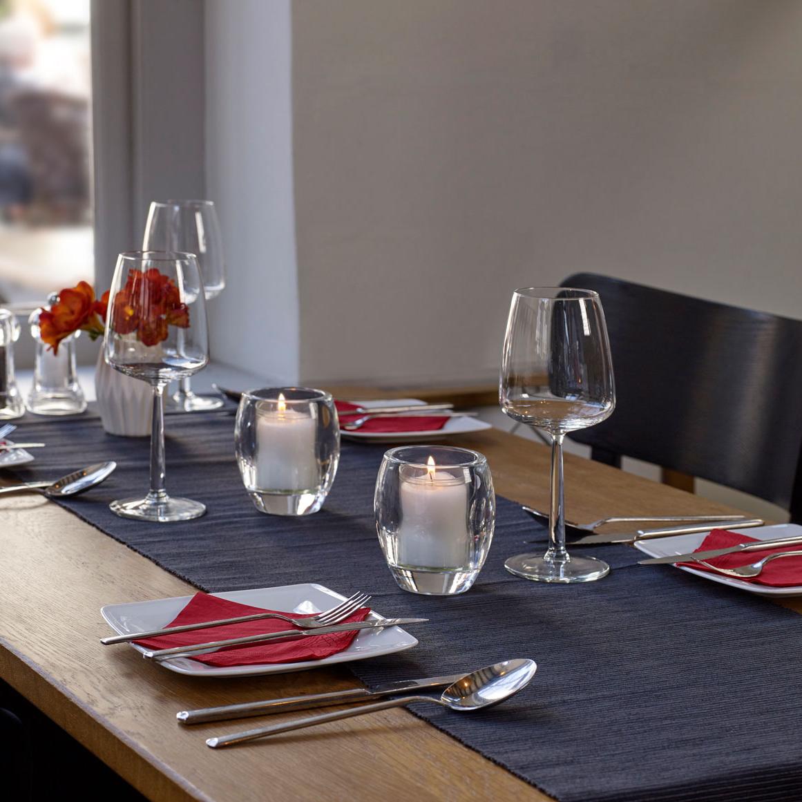 Elegante Tischdeko in einem Restaurant. Anspruchsvoll gestaltet mit weißen Tellern und roten Servietten. Auf dem Tisch steht eine weiße Vase mit roten Blumen und leere Weingläser, begleitet von zwei brennenden Relight Kerzen in einem ovalen Glashalter, die eine gemütliche und einladende Stimmung verbreiten.