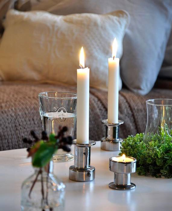 Zwei brennende Tafelkerzen in eleganten, silbernen Kerzenhaltern auf einem weißen Tisch. Ein Wasserglas, eine kleine Glasvase und ein grüner Frühlingskranz liegen daneben. Im Hintergrund sieht man ein gemütliches Sofa mit einer groben Strickdecke und Kissen in braun und creme.