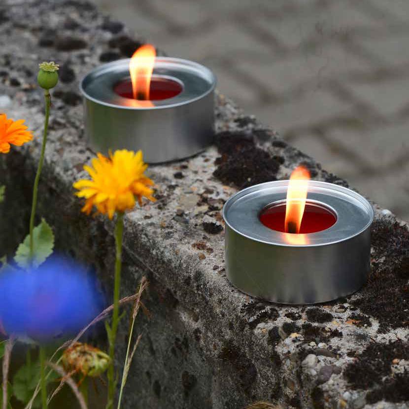 Zwei brennende Marschall-Lichter in einem silbernen Becher mit großer Flamme, auf einer kleinen Mauer, um die sich bunte Blüten ranken.