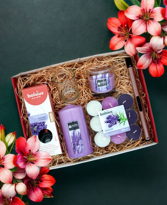Eine mit Stroh ausgelegte Geschenkbox, in der verschiedene Duftkerzen in lila drapiert wurden: eine Lavendel-Stumpenkerze, eine Packung Duftteelichter, ein Duftkerzenglas, ein Raumduft Diffuser sowie ein Teelichtglas und ein rosa Stabfeuerzeug. Die Box steht auf einer dunkelgrünen, ledernen Unterlage, zwischen zwei Blumensträußen mit großen, rosaroten Blüten.