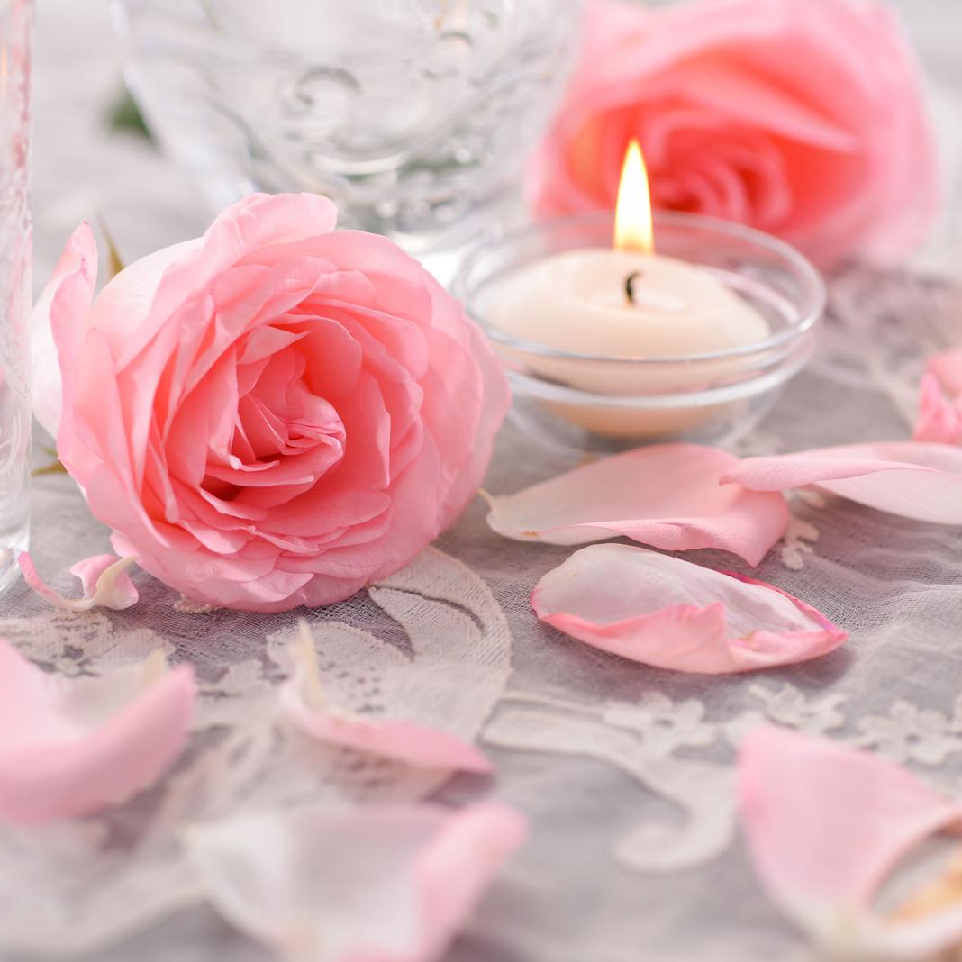 Nahaufnahme eines Tisches, der mit einer verspielten weißen Stick-Tischdecke dekoriert wurde. Darauf stehen mit zarten Ornamenten verzierte Wasserkaraffen aus Glas, Rosen und Blüten in zartem Rosa und romantische Kerzen in Form einer elfenbeinfarbenen Schwimmkerze in einer kleinen Glasschale.