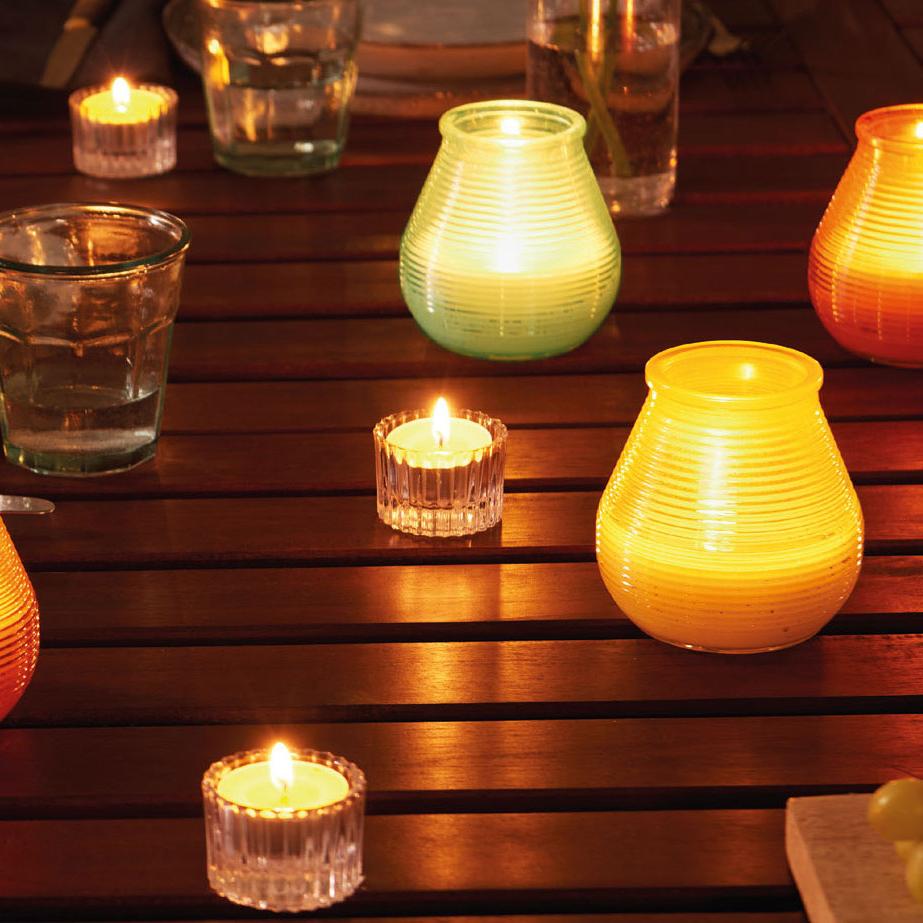 Ein Gartentisch aus dunklem Holz, auf dem neben Tongeschirr und Wassergläsern viele brennende Citronella Kerzen stehen. Einige Patio Windlichter in Orange, Grün und Gelb, sowie ein paar Citronella Teelichte in runden Glashaltern.