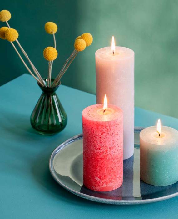 Drei brennende Bolsius Rustik Kerzen in jeweils verschiedenen Größen in Rosa, Pink und Türkis. Die Kerzen stehen auf einem Keramik-Tablett neben einer grünen Vase, die gelbe Blumen enthält, auf einem türkisfarbenen Tisch.