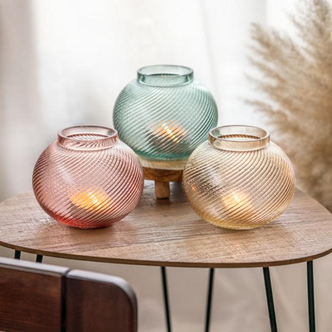 Drei bauchige Kerzenhalter aus buntem Glas, perfekt für deine Frühlingsdeko, stehen auf einem kleinen Holztisch. Im Hintergrund steht eine braune Keramikvase mit buschigem Schilfgras.